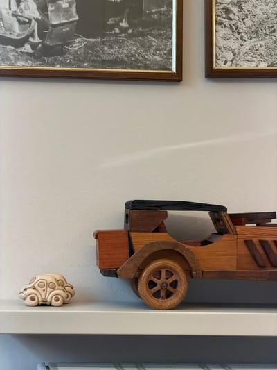 Two framed black and white photographs are mounted on a light-colored wall. Below the photographs, on a white shelf, are two toy cars. To the left is a small, light-colored ceramic car. To the right is a larger wooden toy car with a black roof and wooden wheels. The scene is likely an indoor setting, possibly a home or a display area in Arco, Italy.