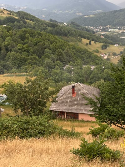The image shows a panoramic view of a mountainous landscape in Mekića Kula, Montenegro. The scene is dominated by lush green forests covering the slopes of the hills, with a small village nestled in the valley below. In the foreground, a rustic house with a grey, corrugated roof and a red chimney stands prominently. The house appears to be in a rural setting, surrounded by dry, golden grass and trees. A few scattered objects, possibly tarps or equipment, are visible near the base of the house. The overall impression is one of tranquility and natural beauty, likely captured during daylight hours under overcast or hazy conditions, given the muted tones of the distant mountains. The composition suggests a high vantage point, offering a sweeping vista of the surrounding terrain.