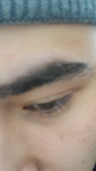 The scene, set in the city of Chinabad, Uzbekistan, captures a close-up, out-of-focus view of a person's left eye and a portion of the adjacent facial skin. The eye is dark and appears partially closed or looking downwards, with visible dark eyelashes. A slight reddish mark is present on the lower eyelid near the inner canthus. A thick, dark eyebrow arches above the eye. The skin is light-toned, showing minor, indistinct blemishes on the cheek area. The upper edge of a gray, ribbed knit beanie or similar headwear is visible at the top of the frame. No discernible actions or interactions are depicted within this static close-up.