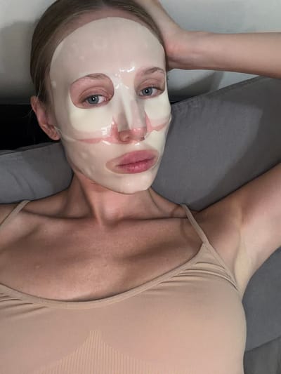 The image shows a close-up, overhead view of a person lying on their back. The person is wearing a sheet mask on their face, which covers their nose and mouth but leaves their eyes and forehead exposed. Their eyes are open and looking upwards, and their hair is visible around the edges of the mask. The person is also wearing a light-colored, sleeveless top that exposes their shoulders and collarbone. Their skin appears fair.

The background consists of a textured gray surface, possibly a sofa or bed. To the left of the person's head, a portion of another limb, likely an arm or leg, is visible, resting on the gray surface. The lighting suggests an indoor setting, with soft illumination that highlights the sheen of the face mask. There are no visible text elements in the image. The overall impression is one of personal care or relaxation.