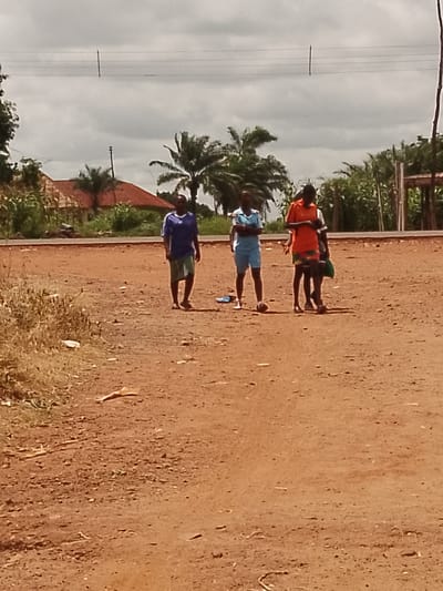 The image depicts an outdoor scene in Adikpo, Nigeria, featuring three individuals standing on a dirt road with sparse vegetation in the background. The ground is a reddish-brown dirt, with some dry grass and scattered debris. In the distance, a building with a red roof and palm trees are visible. Overhead, power lines are strung between poles.
The individuals appear to be young people. The person on the left is wearing a blue t-shirt and a light-colored skirt. The person in the middle is wearing a light blue t-shirt and light blue shorts. The person on the right is wearing an orange t-shirt and patterned shorts. They are positioned in a casual stance, with the person on the middle and right facing towards the viewer, and the person on the left partially turned. There are no obvious interactions between the individuals; they appear to be standing together in a group. The lighting suggests it is daytime, possibly overcast given the cloud cover. The overall environment suggests a rural or semi-rural setting within the city of Adikpo.