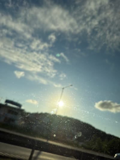 The image is a photograph taken through a dirty windshield, looking out at a bright, sunny sky with scattered clouds. The sun is prominently visible, creating a strong glare and lens flare effect. In the foreground, the windshield has numerous smudges and water spots. Beyond the windshield, a dark, silhouetted hill or mountainside is visible, with some green foliage. To the left, there is a blurred structure that appears to be a building or part of a cityscape, possibly with signage. A street light or pole is also visible on the hill, partially obscured by the sun's glare. The overall impression is of a scenic view from a moving vehicle, likely on a highway, with the sun low in the sky, suggesting either early morning or late afternoon. The location of Mölndal, Sweden, is not discernible from the image itself, as there are no identifying landmarks or contextual clues.
