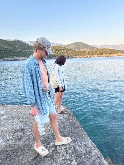 In Dubraljevina, Montenegro, two individuals stand on a concrete pier overlooking a body of water. The person in the foreground, a male with blond hair and a grey baseball cap, wears a blue plaid shirt unbuttoned over a bare chest and light blue shorts. He holds a clear plastic bottle of water in his right hand and has tattoos on his torso and left arm. He is wearing white Crocs. Behind him, a female with dark hair and a white button-down shirt over a black skirt and black sandals looks out at the water. A straw bag is slung over her shoulder. The background features lush, green hills and mountains under a clear blue sky. The water is a vibrant turquoise.