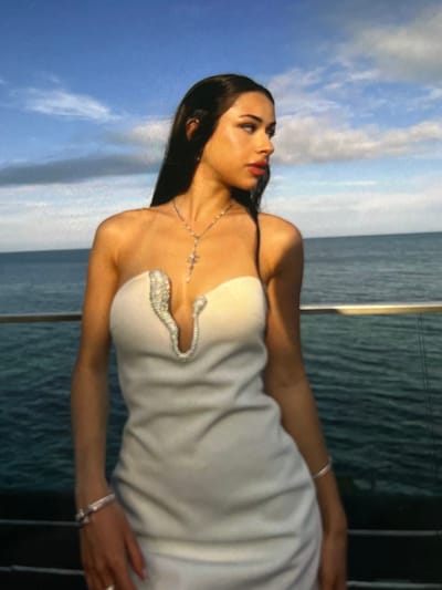 A young woman with dark hair and fair skin stands outdoors, facing right, with the ocean and sky in the background. She wears a white strapless dress with a deep V-neckline embellished with a sparkling, snake-like design. A delicate silver necklace with a cross pendant rests on her décolletage. She also wears a silver bracelet on her left wrist and a ring on her left index finger. The woman's makeup is noticeable, with dark eyeliner and bold lipstick. The water is a deep blue-green, and the sky is a bright blue with scattered white clouds. A metal railing is visible in the foreground, separating the woman from the water. The lighting suggests it is daytime, possibly late afternoon, given the warmth of the light and the angle of the shadows. The scene appears to be on a balcony or deck overlooking the sea. There is no information or visual cues to suggest the image was taken in Korostyshiv, Ukraine.