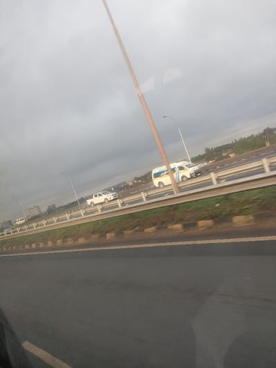 The image depicts a multi-lane highway under an overcast sky, located in Murera ward, Kenya. The perspective is from within a vehicle, looking across and slightly upwards at an adjacent section of the highway. A prominent, slender, light-brown pole traverses the frame diagonally from the upper right to the lower center-left.

In the foreground, a dark asphalt surface, likely a road or shoulder, occupies the bottom third of the frame, showing faint reflections and possibly a yellow road marking on the left. Above this, a sloped embankment with green vegetation and a concrete curb separates the immediate foreground from the main highway.

The midground features a multi-lane highway with traffic flowing from right to left across the frame. Two distinct vehicles are visible: a white commercial passenger van (commonly known as a Matatu in Kenya) with blue markings on its side, and behind it, a white pickup truck. Other indistinct vehicles are present further down the road. The highway is equipped with metal guardrails separating lanes and directions of travel. Several tall, white streetlights with curved arms are spaced along the highway.

The background consists of distant, low-rise buildings and scattered green foliage on slightly elevated terrain, silhouetted against the uniformly grey, overcast sky. No individuals are clearly discernible or identifiable within the scene.