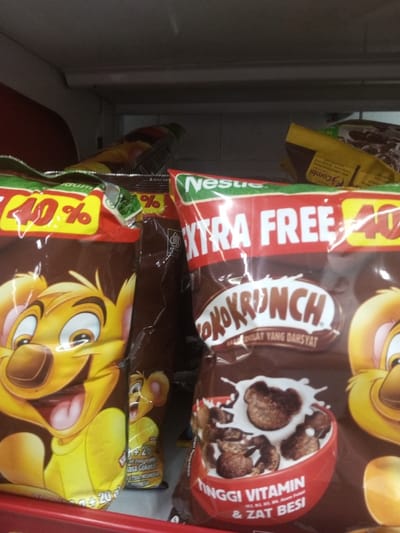 The scene depicts multiple packages of Nestlé Koko Krunch breakfast cereal displayed on a retail shelf in Naung Bo Oe, Myanmar. The foreground is dominated by two large, upright, dark brown plastic bags. The bag on the left features a yellow cartoon bear mascot with a wide smile and outstretched tongue, alongside a "40%" promotional mark. The bag on the right, labeled "Nestlé KOKO KRUNCH" in prominent white lettering, displays "EXTRA FREE" and also a "40%" promotional mark. Below the product name, an image shows chocolate cereal puffs splashing into milk within a red bowl. Additional text on this package reads "TINGGI VITAMIN & ZAT BESI" (High Vitamin & Iron) and "RASAT COKLAT YANG DAHSYAT" (Amazing Chocolate Taste). Other packages are partially visible behind and above the main items, some also bearing similar branding and colors. The environment consists of a light-colored, possibly white or grey, background suggestive of a shelf interior, with a red shelf edge visible at the bottom of the frame. No individuals or specific actions are depicted.