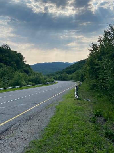 The image depicts a curving asphalt road bordered by lush green vegetation and trees on either side. In the background, rolling hills covered in dense forests recede into the distance, under a dramatic sky with clouds and visible sun rays. The road has clear white lane markings and a yellow line. A silver guardrail runs along the edge of the road. On the right side of the road, near the guardrail, there is some scattered debris. The scene appears to be in a rural or mountainous area, possibly on the outskirts of Zanadvorovka, Russia.