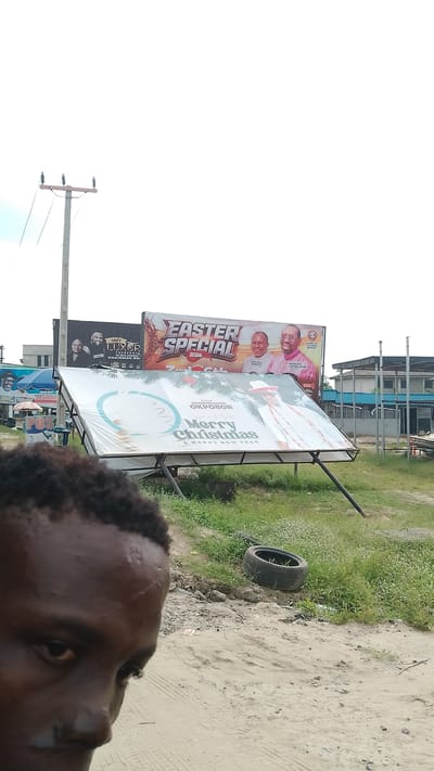 The image shows a partial view of a dark-skinned person in the foreground, with multiple billboards visible in the background. The billboards are displaying various advertisements, including "Easter Special 2026" and "Merry Christmas & Happy New Year." One billboard appears to have fallen or is significantly tilted. The location is indicated by the "Ochenekome Okpobor" billboard which indicates a Warri, Nigeria, location. A tire is discarded on the ground near the fallen billboard. A utility pole is visible in the background, and the general environment suggests a roadside scene.