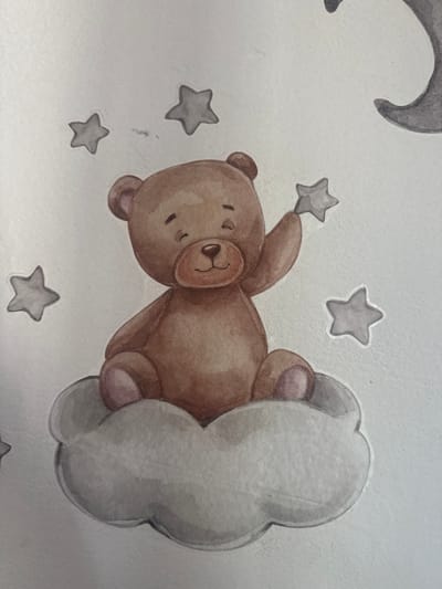 A watercolor painting depicts a brown teddy bear sitting on a grey cloud. The bear is holding a small grey star in its right paw, which is raised. Several other grey stars are scattered around the bear. The background is a pale, textured surface, possibly paper or canvas. The painting has a whimsical, childlike aesthetic. There is no indication of the city of Ushtobe, Kazakhstan, or any other location within the image.