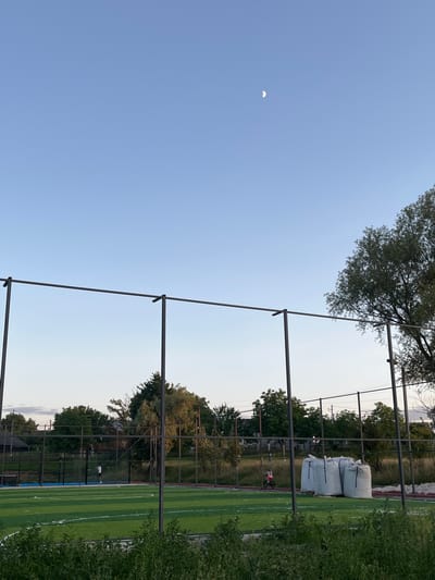 The image depicts an outdoor sports field with artificial turf, likely a soccer or multi-purpose field, in the city of Koroviia, Ukraine. The sky is clear and blue with a crescent moon visible. In the background, there are trees, houses, and power lines. The field is enclosed by a fence, and there are metal poles and horizontal bars that appear to be part of a sports structure or netting system. In the foreground, there is lush green foliage. On the field, a person in white is standing near a blue area, and a person on a bicycle is riding past several large white sacks stacked on pallets. The overall scene suggests a recreational area in a suburban or semi-rural setting.