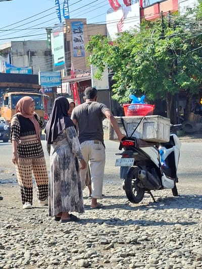 In Blangmetimu, Indonesia, on a sunny day, several individuals are gathered near a street. A man, wearing a grey t-shirt and light-colored pants, stands with his hand on the back of a parked scooter. The scooter is white and has a license plate that reads "BL 5764 ZAO." On the rear of the scooter, a large white cooler box is secured, topped with a red basket containing blue plastic bags. To the left of the man and scooter, two women are conversing. One woman, wearing a pink hijab and a patterned outfit with black and beige circles, appears to be speaking animatedly. The other woman, dressed in a long grey and black patterned dress and a black hijab, faces away from the camera. In the background, a yellow truck and various buildings with signs are visible. The ground is covered with small stones.