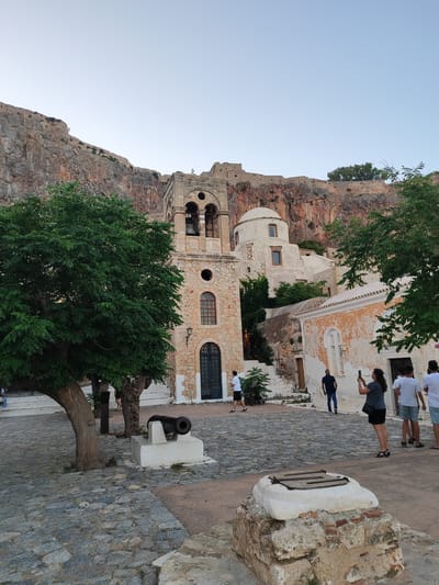 The image shows a view of a stone church with a bell tower and dome, situated against a rocky cliff face. In the foreground, there is a cobblestone courtyard with a small cannon on a pedestal. Several people are present in the courtyard; one woman is taking a photo with her phone, and a group of men are walking away from the camera. Trees with green foliage are visible, framing the church and courtyard. The sky is clear and pale blue. The location is identified as Gefyra, Greece.