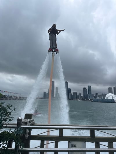 This image depicts a water performance in Qingdao, Shinan District, China, under a heavily overcast sky. Two individuals are engaged in a tandem flyboard maneuver, elevated high above the dark, choppy water by twin powerful jets of water emanating from the flyboard's foot bindings. The primary operator, wearing light-colored trousers and a dark top, stands on the board while supporting a secondary individual, who is positioned in front and extends their left arm horizontally. A reddish-brown pole or hose extends vertically from the flyboard assembly. In the mid-ground, a red personal watercraft, likely powering the flyboard, is partially visible. The background showcases the modern skyline of Qingdao, featuring numerous high-rise buildings and a distinctive white dome structure on the right. To the left, a pier or waterfront area is visible, adorned with a long line of diverse flags. The foreground includes a wooden railing with metal bars and a sign bearing Asian characters, alongside some green foliage.