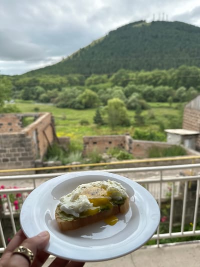 A close-up shot shows a hand holding a white plate with avocado toast topped with a poached egg. The toast is on a slice of bread, with green avocado spread on top, and a runny poached egg with its yolk spilling over. In the background, a mountainous landscape is visible under a cloudy sky. The foreground includes a white railing and some pink flowers. The background also shows some dilapidated structures. The image appears to be taken from a balcony or elevated viewpoint overlooking a rural area with signs of both natural beauty and urban decay. The location is identified as Bavra, Georgia.