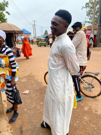 In Barakin, Nigeria, a group of people are gathered outdoors, possibly at a market or along a roadside. The scene is filled with activity and a sense of community. In the foreground, a young man wearing a white robe is smiling and looking down, his arms crossed. Beside him, a person in a colorful striped sweater and a black and white striped robe is lying down, holding a black plastic bag.
Further back, another person wearing a yellow headwrap and a light-colored robe is seen laughing. A bicycle with a partially visible wheel is parked nearby. In the background, various stalls and structures suggest a market environment. A bright orange garment stands out against the muted tones of the ground and buildings. A tuk-tuk, a common mode of transport in Nigeria, is visible, indicating a busy, bustling atmosphere.
The ground is a dusty, earthy color, typical of an outdoor setting in a warm climate. The sky is overcast, suggesting it might be a cloudy day, but the overall lighting indicates it is daytime. The people appear relaxed and happy, engaged in conversation or simply enjoying the moment. The presence of multiple people in traditional attire and the vibrant colors of the clothing add to the cultural richness of the scene. The overall impression is one of everyday life unfolding in a Nigerian community.