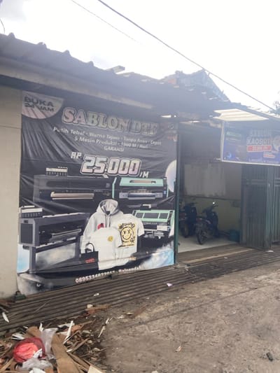 The image shows a storefront with a large banner advertising "Sablon DTF" printing services in Baleendah, Indonesia. The banner features a large price of "RP 25.000" for printing, and showcases various printing equipment, including three large printers. The banner also advertises "Putih Tebal - Warna Tajam - Tanpa Antri - Cepat - 5 Mesin Produksi - 1000 M/Hari - Garansi."  The storefront is partially obscured by the banner and there is a sign on the right side of the image advertising "Sablon Kaos DTF" and prices. Two motorcycles are parked on the left side of the storefront, and a doorway leads into the shop. There is a pile of debris in the foreground of the image, directly in front of the storefront. The location of the storefront is identified as Baleendah, Indonesia with the coordinates -7.005 latitude and 107.631 longitude. The sky is partly cloudy and the ground is a mixture of concrete and dirt.  There are no people visible in the image.
