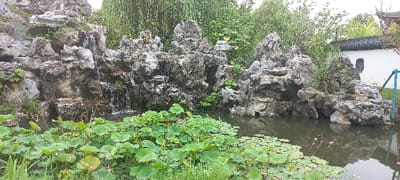 The image shows a man-made rock formation and pond in a garden at the coordinates 30.484, 120.286 in Hangzhou, China. The rock formation simulates a waterfall and a natural mountain range. The pond is filled with lily pads and a small stream of water flows from the rock formation. The pond is surrounded by vegetation and a white building is partially visible in the upper right corner of the image. There are no people visible in the image. The scene is peaceful and serene.