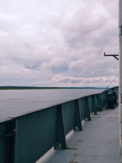 The image depicts a view from the deck of a vessel on a wide river, likely the Yenisei River, near Zotino, Russia. The sky is overcast with a thick layer of grey clouds. The river is calm and extends to the horizon where a dense, green, treeline marks the distant bank. The foreground of the image is dominated by the grey metal railing and deck of the vessel. The railing is robust, featuring large bolts and support structures, suggesting a functional, industrial design. Two large, black bollards are visible on the deck near the railing. The deck itself is a textured metal surface with a pattern of small circular indentations. A thin rope is attached to the railing and extends upwards towards the right side of the frame, where a vertical grey pole is partially visible. The overall impression is one of a utilitarian journey through a natural, yet somewhat somber, landscape under a cloudy sky.