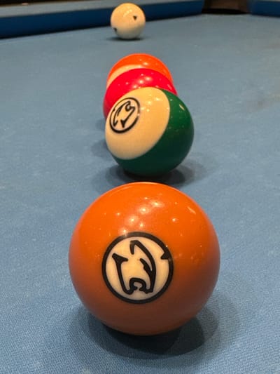 The image shows a close-up of a billiard table. Five billiard balls are visible in a row. The first ball is white. Then there is an orange ball followed by a red ball. Next, there is a green and cream colored ball, and finally an orange ball with a graphic logo on it. The table is covered in light blue cloth. There is no evidence to indicate a location in the city of Bellville, South Africa, or to suggest the presence or actions of any people.