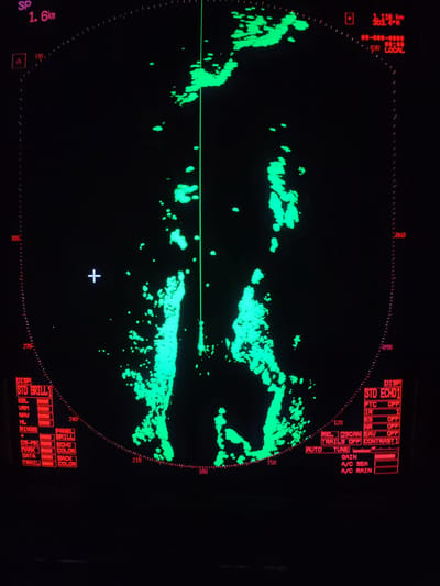 An illuminated radar display, presented in green on a black background, shows radar echoes as bright green irregular blobs across the screen. The display features a circular range with dashed rings and bearing markers (e.g., 330, 300, 270, 060, 090). A prominent vertical green line represents the radar's sweep. A white crosshair cursor is visible slightly left of the center.
Various red digital readouts and setting indicators are present around the perimeter. In the top left, "SP 1.6km" indicates the range scale. The top right shows "1.118 km 303.4°R" likely providing distance and bearing to a target, along with time information such as "030 LOCAL".
At the bottom left, settings related to display brilliance, electronic bearing lines, variable range markers, navigation, heading line, range rings, and target marking are listed under "DISP STD BRILL1". Options for "PANEL BRILL", "ECHO COLOR", and "BACK COLOR" are also shown. On the bottom right, echo processing settings are displayed under "DISP STD ECHO1", including "FTC OFF", "IR 1", "ES OFF", "NR OFF", "REL", "2SCAN", "EAV OFF", "TRAILS OFF", and "CONTRAST 1". Manual adjustments for "AUTO TUNE", "GAIN", "A/C SEA" (Anti-Clutter Sea), and "A/C RAIN" (Anti-Clutter Rain) are also visible with associated bars, suggesting a marine or similar radar system operating in variable conditions.
The overall image depicts an active radar display, likely within a vessel's control room, used for navigation or target detection. No people are visible.