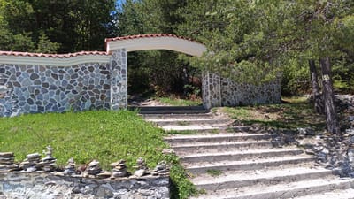 A stone archway leads up a series of concrete steps in Sini Vrah, Bulgaria. The archway is constructed of rough-hewn stone with a red-tiled roof. To the left of the stairs, small stacks of flat stones are piled on top of a stone wall. Green grass and trees surround the area.