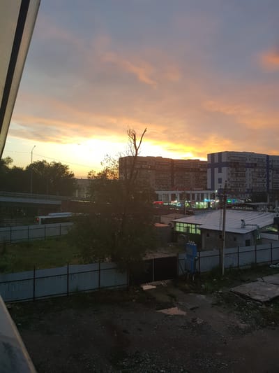 The image depicts a late evening scene in the city of Бұлақты, Kazakhstan. A sunset paints the sky with hues of orange and pink, casting a warm glow over the urban landscape. In the foreground, a dark, gravelly area with puddles suggests recent rain. A corrugated metal fence runs horizontally across the frame, separating the foreground from a grassy area beyond.
Further back, a tree with bare branches stands prominently. Behind the tree, a road or elevated walkway is visible, with a white bus driving on it. Numerous apartment buildings and commercial structures form the city skyline. Some of these buildings have illuminated signs, including one that reads "TOIMART." Power lines and a street lamp are also present. The overall impression is of a residential and commercial area at dusk, characterized by its modest architecture and the natural beauty of the sunset.