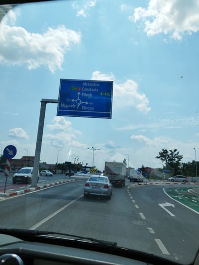 The image shows a view from inside a vehicle on a sunny day, looking forward towards a highway intersection and a large blue directional sign. The sign indicates routes to Alexandria, Constanta, Pitesti, Măgurele, and Clinceni. Several cars and trucks are visible on the road, as well as traffic signs and streetlights. The environment appears to be a suburban or semi-urban area in Bragadiru, Romania, with buildings and trees lining the roadside. The sky is blue with scattered white clouds.