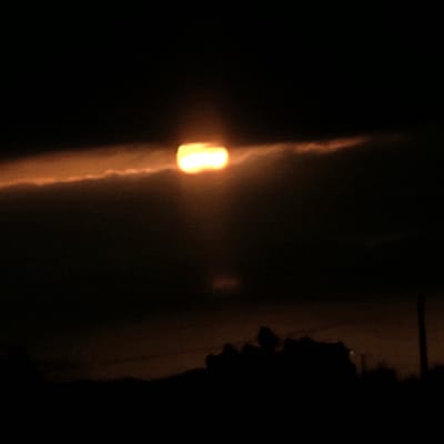 This image depicts a sunset or sunrise over Nzalat Laadam, Morocco. The scene is dominated by a dark, cloudy sky. A bright, horizontally elongated, orange-yellow celestial body, consistent with a sun obscured by atmospheric conditions, is centrally positioned in the upper portion of the frame. This light source is visible through a distinct, narrow horizontal band of clouds, which are illuminated with an orange glow immediately surrounding the light. Below this, the lower half of the image features a dark, indistinct silhouette of what appears to be a landscape or urban structures against the horizon. On the far right, a vertical dark shape, possibly a utility pole, is discernible against the darkened sky. No individuals, specific actions, or interactions are visible in the scene.