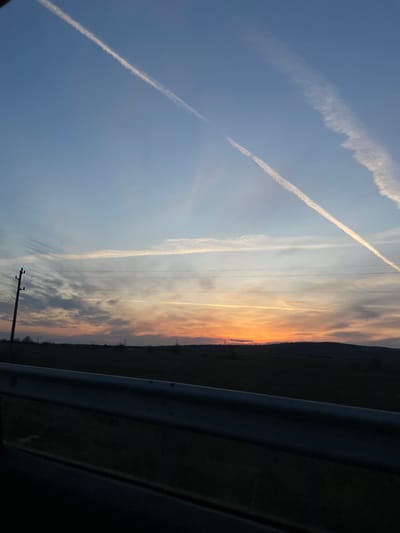 An outdoor scene depicts a sunset over a rural landscape in Gorna Mahala, Bulgaria. The upper sky is clear blue, transitioning to vibrant orange and yellow hues along the horizon. Multiple white contrails streak across the sky; two prominent ones cross diagonally from the upper left and upper right. Other faint, elongated cloud formations are also present horizontally in the mid-sky. Below the horizon, a dark, expansive rural landscape is visible, consisting of fields and distant, low-lying hills or a dense treeline. A dark utility pole with multiple horizontal wires extends vertically on the left side of the mid-ground. A metallic guardrail occupies the lower portion of the frame, indicating the viewpoint is from a vehicle or roadside. No individuals or significant human activity are depicted within the scene.