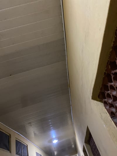 The image shows the interior of a room in Sabo, Nigeria. The walls are constructed of vertical panels and are painted in a light, possibly beige, color. At the top of the frame, parts of the ceiling are visible, including areas with decorative brown elements. There are windows on the right side of the room. The lighting appears to be provided by a bright light source reflected on the wall. There are no people visible in the image.