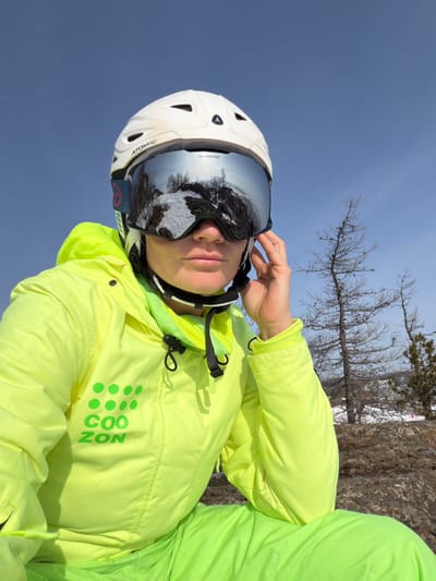 An adult female individual is depicted in an outdoor, elevated winter environment in Zelenaya Polyana, Russia. She is positioned facing the camera, wearing a white "ATOMIC" brand ski helmet and large, mirrored "GLASSLADE" brand ski goggles. The goggles reflect a snowy, rocky, forested landscape under a clear blue sky. She is dressed in a neon yellow/lime green ski jacket with a hood, featuring a dark green "COO ZON" logo and dot pattern on the left chest, and matching ski pants. Her left hand is raised to the right side of her head, with fingers near the temple area, seemingly adjusting the goggle strap. The background consists of sparse, bare trees and rocky ground under a clear blue sky.