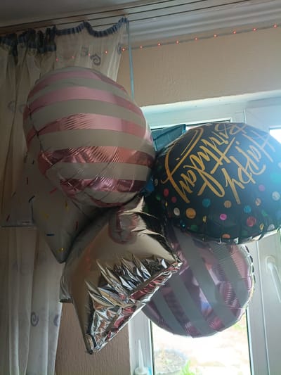 In an indoor setting, likely a residential room in Litynia, Ukraine, a cluster of five balloons is suspended from above. The balloons are of various shapes and designs. One large, round balloon is black with gold lettering that reads "Happy Birthday" and is decorated with colorful polka dots. Another round balloon features pink and grey stripes. A star-shaped balloon is silver with a crinkled, metallic finish, and another star-shaped balloon is light grey with a pattern of colorful sprinkles. A fifth balloon, partially obscured, also appears to have pink and grey stripes. The balloons are hanging in front of a sheer, white curtain with blue trim, and a window with a white frame and handle is visible in the background, suggesting the image was taken near a window during daylight hours. A string of small red LED lights is visible along the ceiling line. No people are present in the frame.