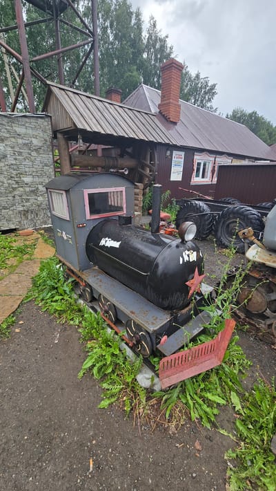 A small, black, metal, toy train sits on a track outside a building. The train has a red star on its front and white writing on its side. It is surrounded by green weeds and a dirt path. In the background, there is a building with a red roof and brick chimney, as well as a metal structure and trees. The sky is overcast. This scene is located in Bol'shoi Mashlyak, Russia.