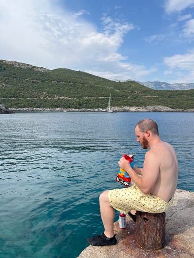 A shirtless man with a beard sits on a rustic wooden stump on a concrete pier, holding a bag of Monte Chips. He is wearing light yellow shorts with a pineapple pattern and black slip-on shoes. To his right, a red and silver can rests on the pier. The sea in front of him is a vibrant turquoise, with gentle ripples reflecting the blue sky and scattered white clouds. In the distance, lush green hills rise from the coastline, dotted with trees. A white sailboat is anchored in the bay. The overall scene suggests a relaxed moment by the water in a scenic, coastal environment, likely near Dubraljevina, Montenegro, given the picturesque mountainous landscape and clear blue sea.