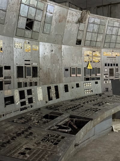 This image depicts an abandoned and severely deteriorated control room, characteristic of an industrial facility, specifically resembling the control rooms found within the Chernobyl Nuclear Power Plant, located in the Chernobyl Exclusion Zone, north of Kyiv, Ukraine. No people are present.

The foreground features a large, curved control console, dark gray and heavily encrusted with grime and dust. Numerous square and circular cutouts reveal missing buttons, switches, and panels, leaving empty cavities and exposed wiring. Debris, including what appears to be organic material and discarded items, is visible within these hollowed-out sections.

Behind the console, a multi-tiered wall of vertical panels, lighter gray and extensively corroded, displays peeling paint and heavy staining. The lower and middle tiers exhibit empty rectangular slots where instruments once resided, alongside faded yellow-on-black labels and rows of small, dark, possibly non-functional indicators. A prominent yellow triangular radiation hazard symbol with a black trefoil is affixed to a panel on the middle tier, slightly right of center. The upper tier consists of multiple shattered or missing display windows, revealing dark interiors and underlying structural elements. Loose wires hang from various panels, and a piece of light-colored fabric or plastic dangles from the ceiling near the top center.

The scene portrays significant neglect and advanced decay, indicative of prolonged abandonment in an unpowered, dimly lit environment.