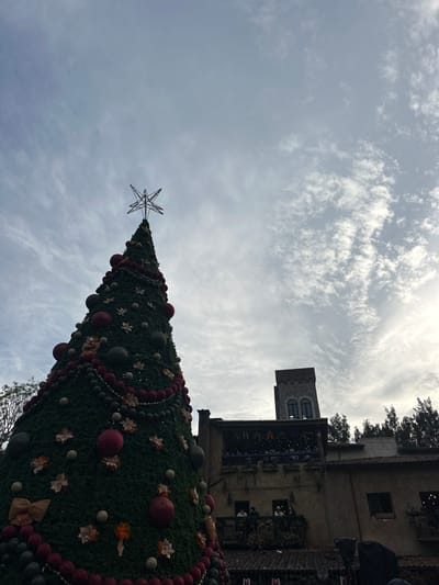 A tall Christmas tree with a star on top stands prominently against a cloudy sky. The tree is adorned with red and gray ornaments, as well as garlands of red and white beads. Behind the tree, a stone building with a tower and balconies is partially visible, suggesting an urban or European setting. The image is taken from a low angle, emphasizing the tree's height and the imposing architecture.