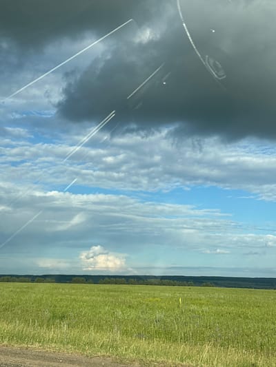 The image depicts a vast, sun-drenched green field, likely a meadow or pasture, under a dynamic sky. Numerous white contrails crisscross the sky, indicating recent airplane activity. These trails are superimposed on a backdrop of fluffy white clouds interspersed with patches of clear blue sky. Dominating the upper portion of the frame are darker, more ominous clouds, hinting at potential weather changes. In the distance, a line of dark green trees forms a horizontal band, possibly marking the edge of the field or a forest. A single, brightly lit white cloud stands out against the darker sky on the horizon, and a very subtle rainbow arc is visible in the distance. The foreground of the image shows a dirt road or path. The overall impression is a natural landscape under a dramatic sky, viewed from an elevated or slightly angled perspective, possibly through a window or windshield, given the subtle reflections or smudges visible in the top portion of the image. The location in Tatarstan, Russia, suggests a rural or semi-rural setting.