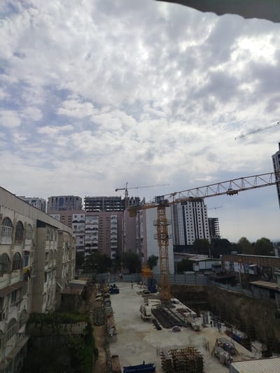 The image depicts a construction site in an urban environment, likely Khujand, Tajikistan. The scene is dominated by several large apartment buildings in various stages of construction, interspersed with older, occupied buildings. Multiple construction cranes are visible, with one prominent yellow tower crane in the foreground. The ground is a mix of concrete, dirt, and piles of construction materials such as rebar, wooden planks, and bundles of material wrapped in plastic. A few people are present on the site, seemingly engaged in work.
The time of day appears to be daytime, judging by the ambient light, and the weather is overcast with scattered clouds. The sky is a muted grey, suggesting a cloudy or possibly approaching rain. The older buildings have a distinct architectural style with arched windows. The new buildings show modern construction, with some nearing completion while others are still skeletal structures. Fencing encloses parts of the construction area.
There is some visible text on billboards or posters in the background, but it is too small and blurry to read. The overall impression is one of ongoing development and urban expansion.