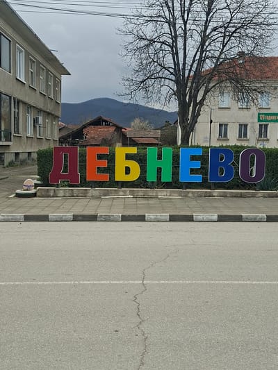 The image shows a street scene in Debnevo, Bulgaria. Large, vertically-oriented, colorful letters spelling "ДЕБНЕВО" (Debnevo in Cyrillic) stand along the sidewalk. The letters are red, orange, yellow, green, light blue, dark blue, and purple. The sidewalk is lined with concrete bumpers. The road surface is gray asphalt. Buildings flank either side of the street. A bare tree is visible in the background. Mountains are visible in the distance, under an overcast sky. A sign reading "120 ГОДИНИ Ч" is on one building. There's a decorative planter next to the town name sign.