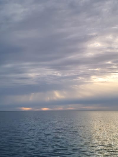 The image depicts a seascape under a cloudy sky. The water is calm, with gentle ripples across the surface. The sky is mostly covered with grey clouds, but there are breaks in the clouds near the horizon on the right side, allowing sunlight to stream through. This creates shafts of light that illuminate the water in patches. A sliver of orange is visible along the horizon on the left. A small bird is flying over the water in the center. The location is Shcheleiki, Russia.