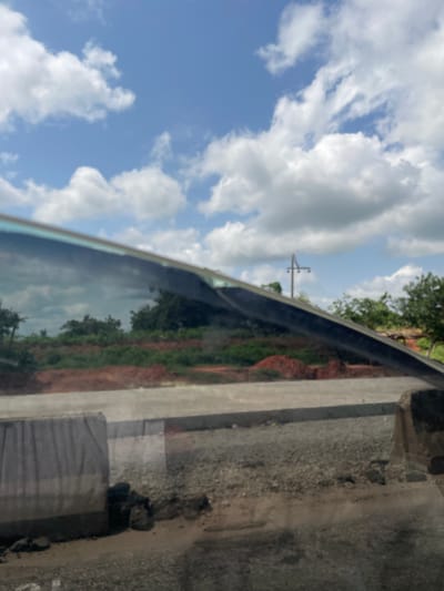 The image depicts a reflective surface, likely a car window, showing a mirrored view of a landscape. The foreground is dominated by the dark, glossy surface, hinting at the presence of a vehicle. Reflected in this surface are elements of the environment outside.

The reflected scene includes a bright blue sky dotted with fluffy white clouds, suggesting it is daytime and likely fair weather. Below the sky, a green landscape is visible, with trees and vegetation indicating a natural or rural setting. There are also hints of red earth, possibly a dirt road or exposed soil. In the distance, a utility pole with wires is faintly visible, a common sight in many populated areas.

To the right of the reflective surface, the physical environment is more clearly visible. This includes a rough, grey textured surface, possibly concrete or asphalt, with scattered gravel. A wooden plank or beam is also visible, situated on top of this rough surface. The overall impression is of an outdoor scene, perhaps taken from a roadside or a similar vantage point. The image is oriented vertically, with the sky and landscape appearing at the top, and the physical surroundings at the bottom. No people or specific activities are discernible in the image.
