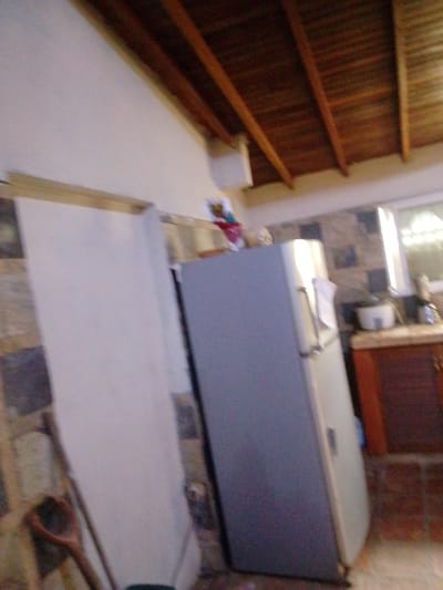 The image depicts an interior view of a kitchen in Atamo Sur, Venezuela. The scene features a refrigerator, a wooden ceiling with exposed beams, and a countertop with appliances. On the left side of the image, a large, white appliance, possibly a refrigerator or freezer, is visible. To its right, a gray refrigerator stands prominently. Above the refrigerators, a wooden ceiling with parallel beams spans the space, suggesting a rustic architectural style. The countertop in the background displays what appears to be a white appliance, likely a blender or food processor, and a small, round object, possibly a bowl or pot. The walls are tiled with a dark and light pattern. The overall impression is of a functional kitchen space.