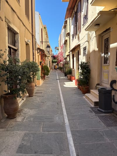 The image depicts a narrow, sun-drenched street in Rethymno, Greece, with traditional architecture and potted plants lining the walkway. The buildings on either side are multi-story structures with cream and ochre colored facades, featuring wooden shutters and balconies. The street is paved with large, grey stone slabs, bisected by a white painted line down the center.
On the left side of the street, large terracotta pots contain lush green plants and trees. Further down the street, more potted plants and greenery are visible, some with vibrant pink flowers. Several people are walking in the distance, casually dressed, suggesting a relaxed, daytime atmosphere.
On the right side of the street, a building with a light-colored facade has a white door with an ornate metal grate and the number "52" above it. A black outdoor trash receptacle with an ashtray is situated on a small set of steps leading up to the doorway. Graffiti is visible on the wall next to the trash can. The overall impression is of a charming, pedestrian-friendly street in an old European town.