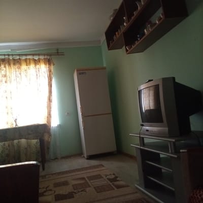 The image shows an interior view of a room, possibly a living room or kitchen, with light green walls. A large, light-colored refrigerator stands against the left wall. In the center of the room, a patterned rug is visible on the floor. To the right, an old-fashioned CRT television is placed on a grey stand. Above the refrigerator and on the right wall, dark brown shelves are mounted, containing various items that appear to be glassware. A window with sheer, patterned curtains is on the left side of the frame, with a table covered in a patterned cloth positioned in front of it. The lighting suggests it is daytime, with light streaming through the window. The overall impression is of a somewhat dated and modestly furnished living space. There is no indication of any specific location in Slobidka, Ukraine, beyond it being an indoor scene.