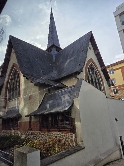 The image displays the exterior of a church building in Levallois-Perret, France, under an overcast sky. The central structure features a dark-colored, steeply pitched roof culminating in a slender spire topped with a cross. The church walls are light beige with reddish-brown trim accentuating large, arched windows containing ornate tracery. Below the main windows, smaller sections of the church facade incorporate reddish-brown brickwork and a lower foundation of textured stone veneer. Two smaller, dark-roofed projections with barred windows or railings extend from the lower portion of the church. In the foreground, a low, light-colored concrete wall and a square concrete post are visible, with green and autumnal-toned shrubs and foliage growing behind the wall. In the background, to the right, a modern, light-colored multi-story building and an older, yellow-orange building with visible windows are partially obscured by the church. Faint white text reflecting on the left edge of the image suggests the photograph was taken through glass. No individuals are present in the scene.