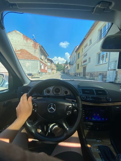 The image is taken from inside a car, looking out through the windshield. The scene depicts a street view in Belogradchik, Bulgaria. The perspective is as if the viewer is the driver, with a hand on the steering wheel visible in the foreground. The steering wheel is black leather with a Mercedes-Benz logo in the center and control buttons. Part of the car's dashboard is also visible, including the car's infotainment screen displaying icons and the time "10:02".
The setting is an urban environment with buildings lining the street. The architecture suggests a European town, with multi-story buildings featuring brick and stucco facades, and visible windows. The sky is bright blue with a few fluffy white clouds, indicating a sunny day. The lighting suggests it is daytime, likely mid-morning.
The activity appears to be driving through a residential or mixed-use area. The orientation of the image, with the buildings and sky appearing to be on the left and the car interior on the right, is unusual, suggesting the photo was taken with the camera rotated.
Notable details include the brickwork on one of the buildings, which shows signs of wear, and the general architectural style of the town. There is no strong emotional content conveyed, it is a straightforward depiction of a driving scene. Text visible includes the Mercedes-Benz logo on the steering wheel and the time on the car's display.
