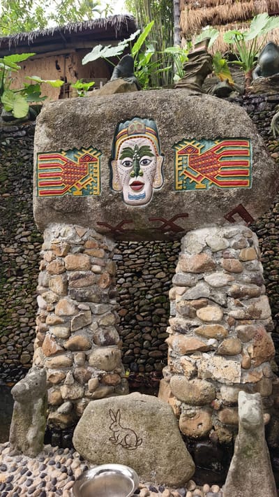 An outdoor scene in Jiyang District, China, depicts a decorative stone archway or monument integrated with natural elements. The central feature is a large, grey horizontal stone lintel supported by two substantial pillars constructed from irregularly shaped, cemented natural stones. The lintel displays three deeply recessed, vibrantly painted carvings: a stylized human face or mask in the center with green, white, red, black, and blue details, flanked by two symmetrical geometric motifs resembling stylized birds or fish, colored primarily in yellow, red, and green. Dark reddish-brown angular patterns are painted on the underside of the lintel between the pillars.
Behind the stone structure, a vertical wall is composed of numerous small, dark, smooth river stones, some exhibiting green moss, indicating the presence of moisture or running water that appears to flow downwards. Lush green vegetation, including large-leafed plants and tall bamboo stalks, densely populates the upper background. Portions of a building with a thatched roof are visible through the foliage. Several dark, rough-hewn stone figures, some appearing as abstract forms and others resembling frogs, are positioned on elevated ledges amidst the greenery.
The immediate foreground consists of a ground surface covered in small, smooth pebbles. Resting on these pebbles is a lighter-colored stone with an etched outline of a rabbit. Adjacent to it, a reflective, oval-shaped metallic basin is visible. Water is present at the base of the stone pillars, collecting in a dark pool. No human figures are present in the scene.