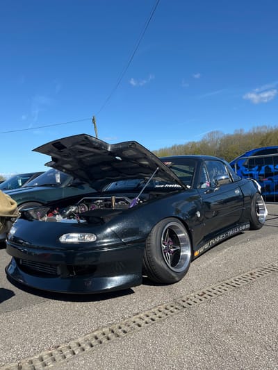The image shows a black Mazda MX-5 parked on an asphalt surface in Silverstone, United Kingdom. The car's hood is open, revealing a modified engine bay with purple accents. A decal reading "www.TUG2S-TAILS.com" is visible on the car's side. A blue vehicle is partially visible behind the Mazda. The sky is blue with scattered clouds. A utility pole and wires are visible above the car. A drain runs alongside the car.