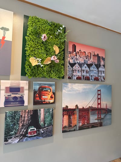 This media file displays a collection of framed photographs and a decorative green wall art mounted on a light gray wall.
The top row features two large landscape orientation photographs. The left image shows a cityscape with a row of Victorian houses in the foreground against a backdrop of urban buildings, bathed in a warm, sunset-like light. The right image is of the Golden Gate Bridge, captured during what appears to be late afternoon or early evening, with the sky showing soft clouds and a warm glow reflecting off the iconic red structure.
Below the cityscape and to the left of the bridge photo is a square green wall art composed of dense, artificial foliage, resembling a moss wall. It is adorned with artificial flowers in shades of pink and white, and two golden, leaf-shaped decorative elements.
The second row contains three smaller photographs arranged horizontally. On the left, an orange Volkswagen Beetle is parked near a body of water, its license plate visible. In the center, a vintage Polaroid land camera is the subject of a close-up shot. To the right of the camera, a photograph shows a red and white vintage car driving through a tunnel carved into a giant tree trunk, likely a redwood tree in California. The scene suggests daytime.
The bottom section of the media file includes a portion of another framed artwork that features a stylized illustration of a tree with some abstract elements.
The overall setting appears to be an interior space, possibly a home or a studio, decorated with these pieces of art. The wall color is neutral, allowing the images to stand out. There are no people visible in any of the photographs. The collection seems to celebrate iconic landmarks, vintage aesthetics, and nature-inspired elements.