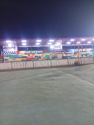 An outdoor basketball court with the text "DAM FOUNDATION BASKETBALL CHAMPION" reflected on the ground. In the background, there's a painted wall with colorful geometric shapes and the phrase "DREAM BIG. PLAY BIGGER". Above the wall, there's a shaded structure with bright lights illuminating the scene at night. Signs with text, including "LIVIC RECREATION COMPLEX" and "DAM 25", are visible on the structure.